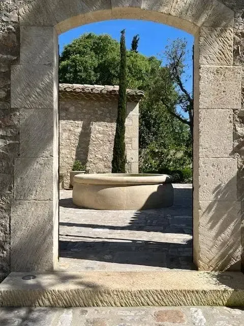 Une ouverture en arche de pierre donnant sur une cour ensoleillée, avec un puits ou une fontaine circulaire en pierre et des arbres au-delà.