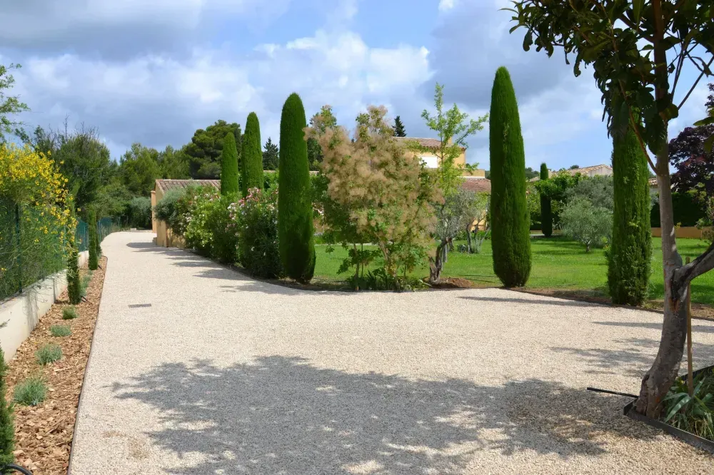 Allée de jardin sinueuse en gravier, bordée de grands cyprès et de verdure sous un ciel nuageux