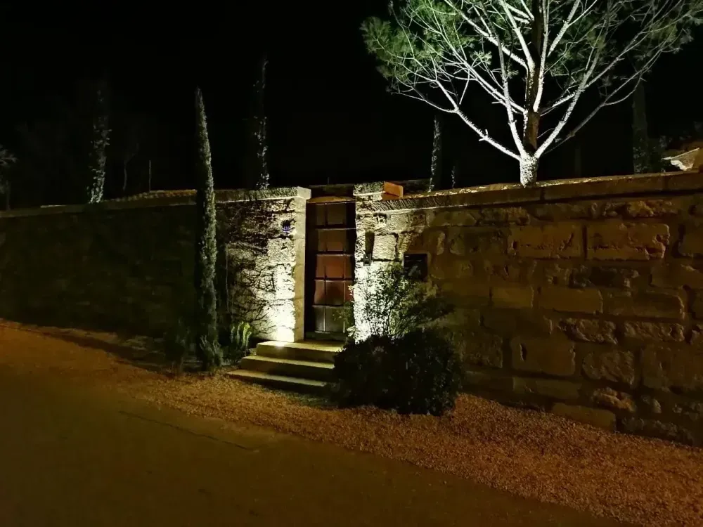 Vue nocturne d’un mur et d’un portail en pierre éclairés par un lampadaire, avec des arbres en arrière-plan