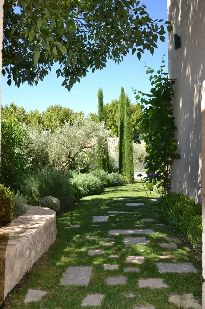 Allée de jardin avec des pas japonais, une végétation luxuriante, de grands cyprès et un mur de pierre ensoleillé.