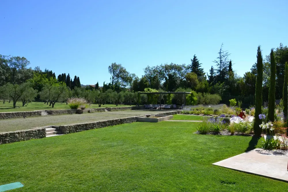 Jardin ensoleillé et paysager avec pelouse verdoyante, marches en pierre, massifs de fleurs et arbres sous un ciel bleu dégagé