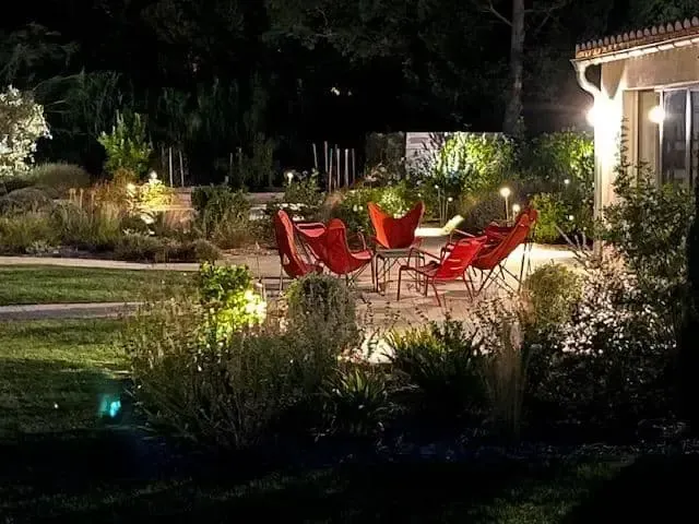 Des chaises de terrasse rouges autour d’une table éclairée dans un jardin la nuit, à côté d’une maison baignée d’une lumière extérieure chaleureuse