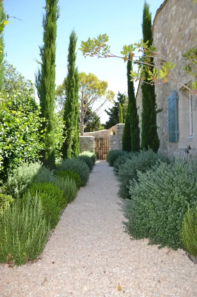 Allée de gravier dans un jardin soigneusement entretenu, à côté d’un bâtiment en pierre, avec de grands cyprès et des arbustes.