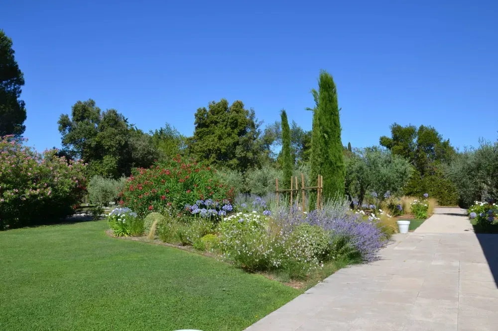 Jardin ensoleillé avec des fleurs colorées, une pelouse verte, de grands cyprès et une allée pavée sous un ciel bleu