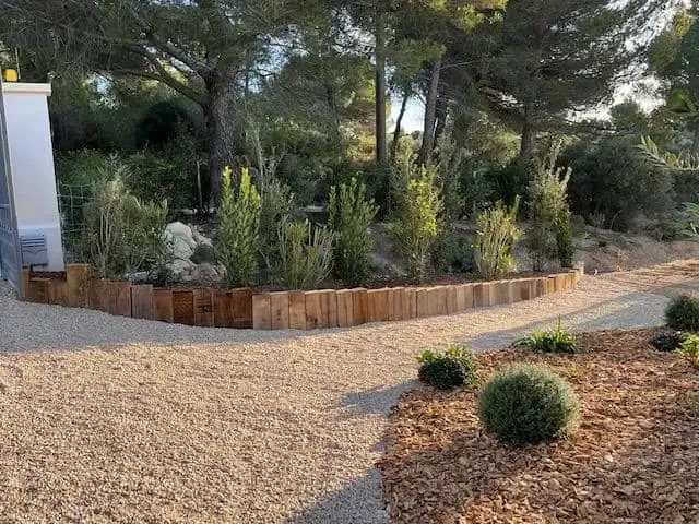 Allée paysagée en gravier bordant une jardinière surélevée en bois, agrémentée d’arbustes et de grands arbres