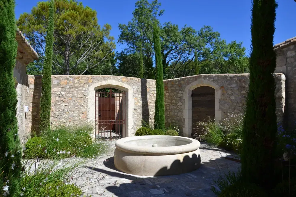 Cour en pierre avec une fontaine circulaire au centre, des murs de pierre en arcades et de grands arbres baignés de soleil