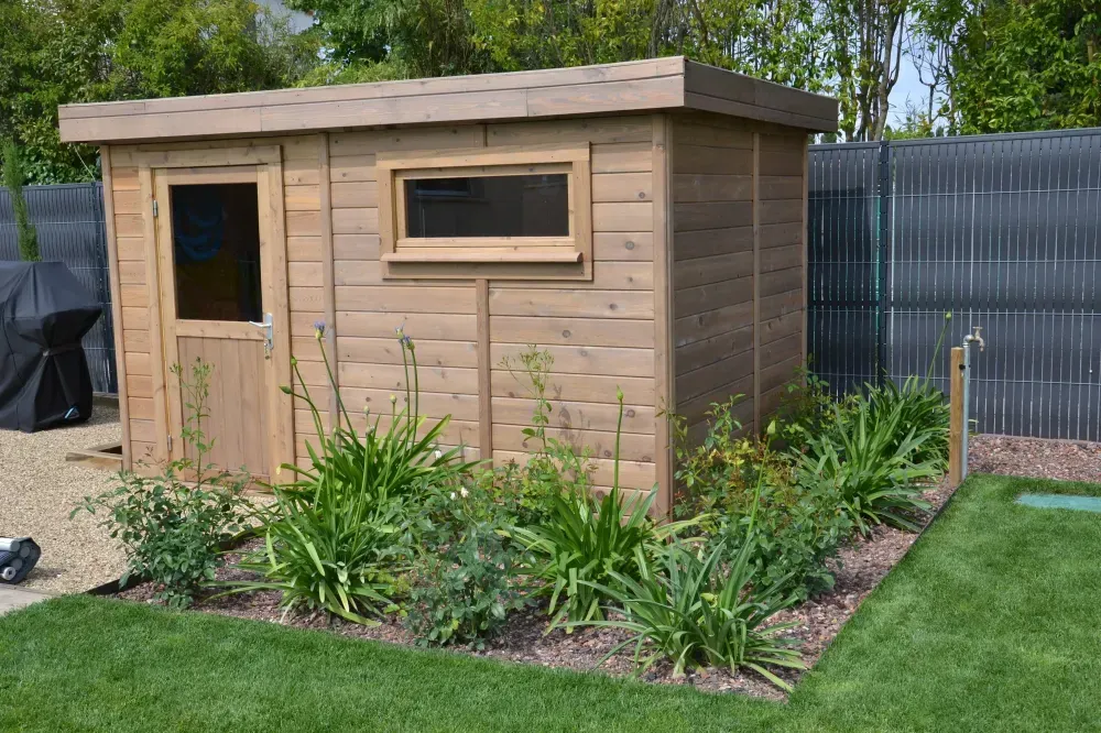 Abri de jardin en bois avec une fenêtre et une porte, entouré de plantes sur une pelouse dans un jardin.