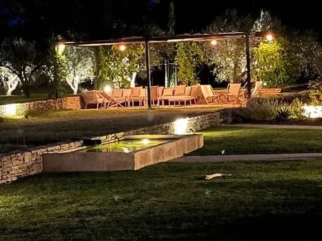 Salon de jardin éclairé avec pergola, sièges et lumières chaleureuses, au bord d’un bassin miroir de nuit