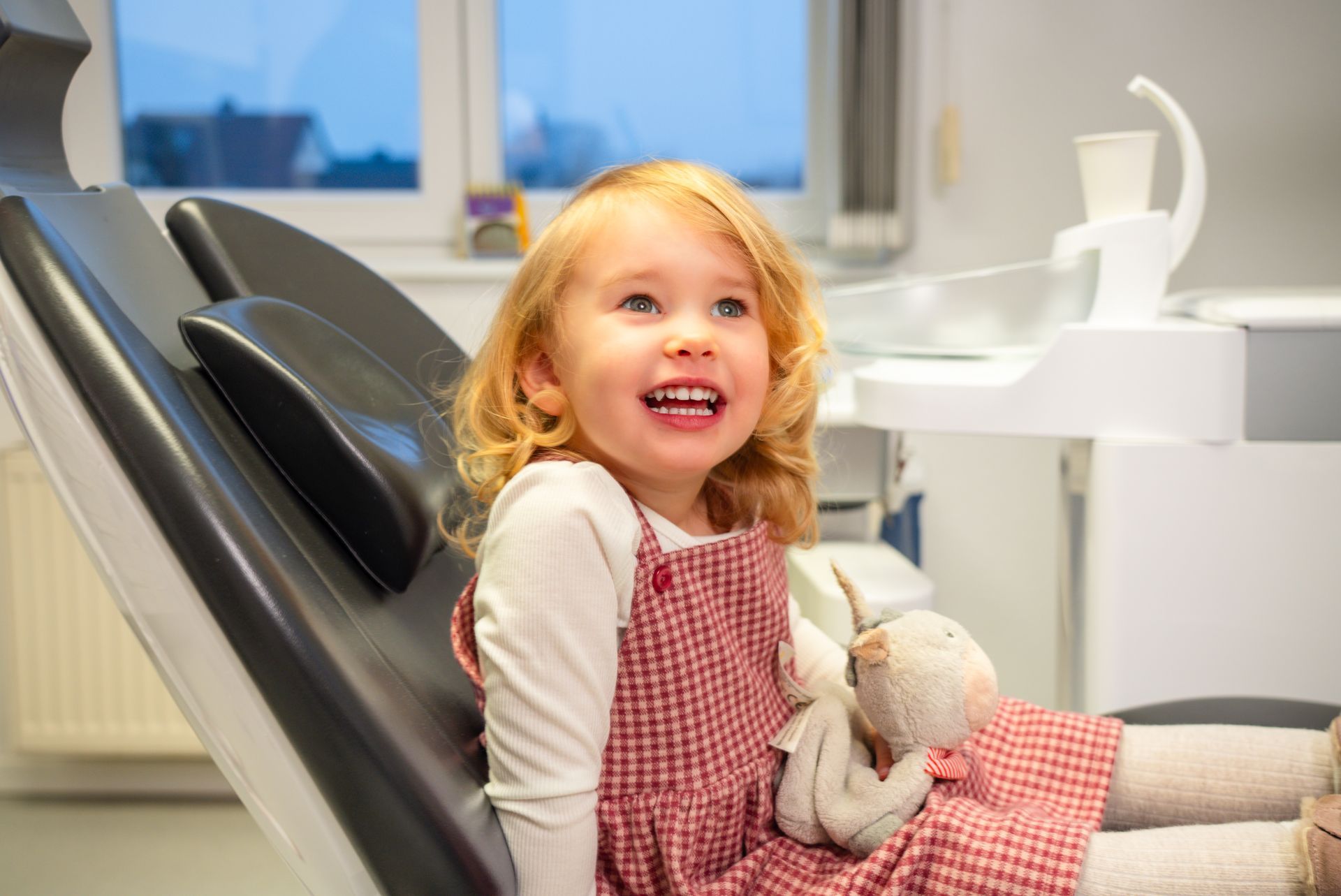 Ein lächelndes Kind, das auf einem Zahnarztstuhl sitzt und ein Plüschtier hält.