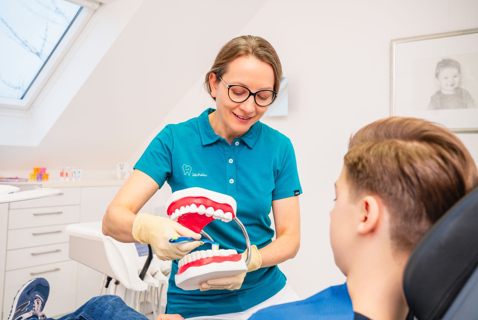 Ein Zahnarzt in einem türkisfarbenen Poloshirt demonstriert einem Patienten anhand eines Zahnmodells die richtige Zahnputztechnik.