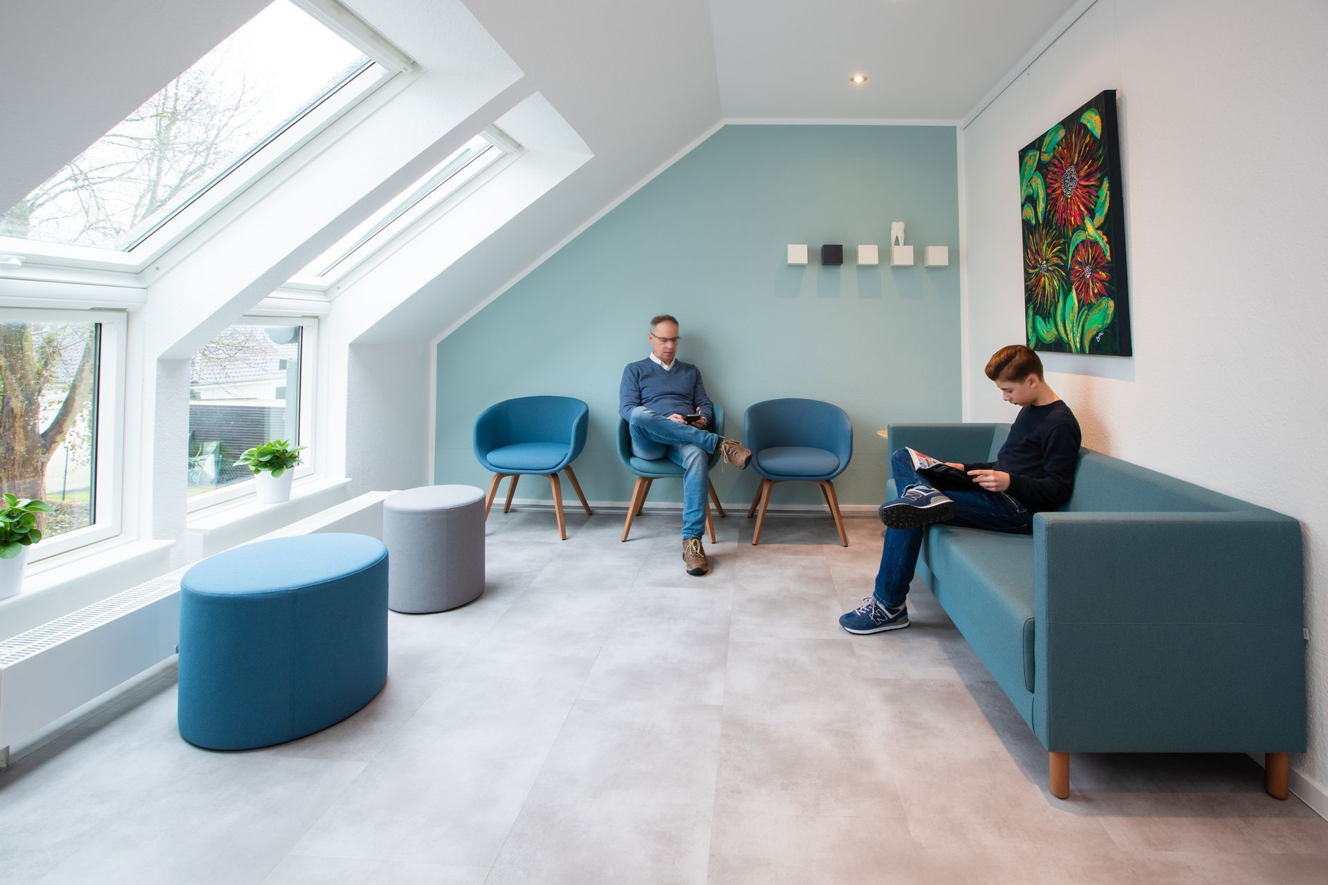 Ein helles, modernes Wartezimmer mit türkisfarbenen Stühlen, einer Couch, einer hellblauen Wand, Oberlichtern und zwei Personen, die schweigend dasitzen.