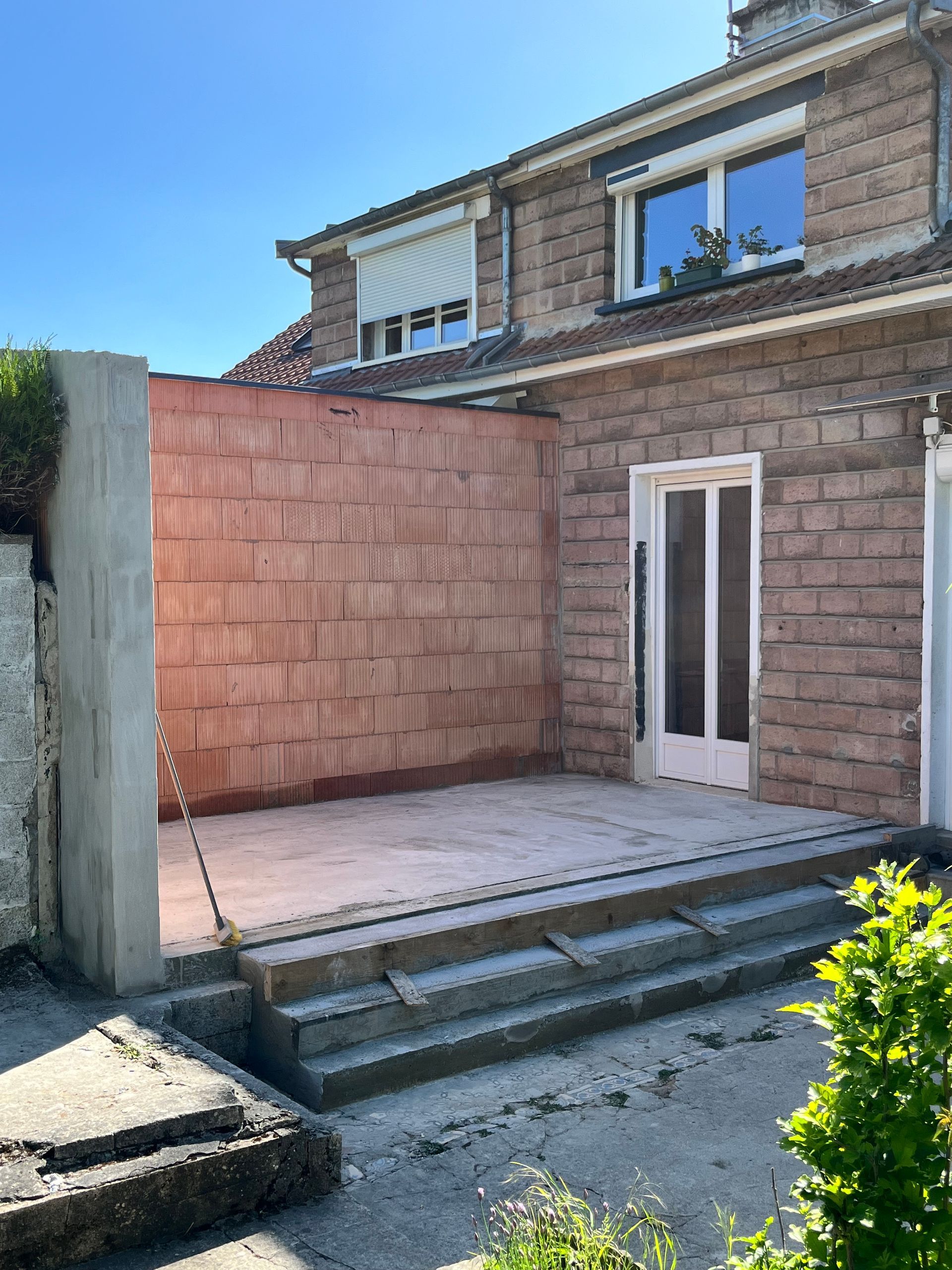 Terrasse extérieure avec marches en béton, murs en briques rougeâtres et en béton. Maison en arrière-plan.