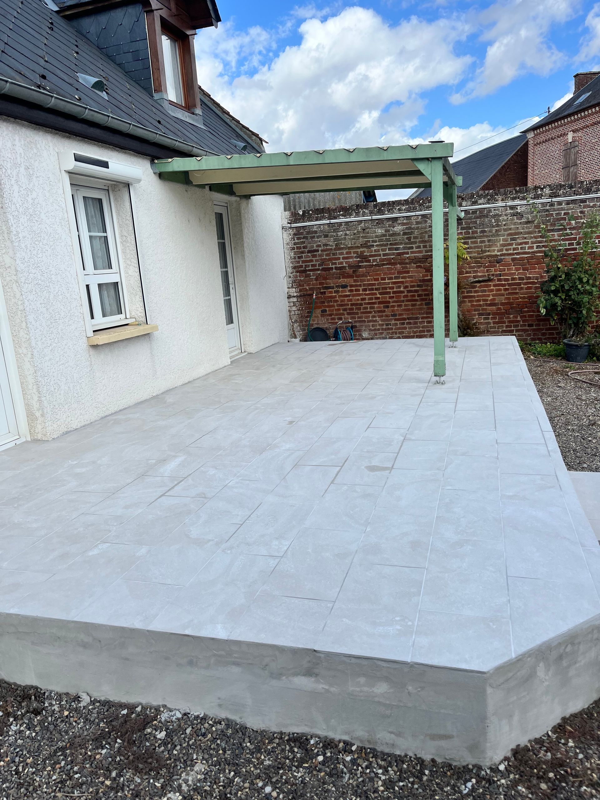 Terrasse en béton avec pergola verte adossée à un bâtiment blanc. Mur de briques rouges en arrière-plan.