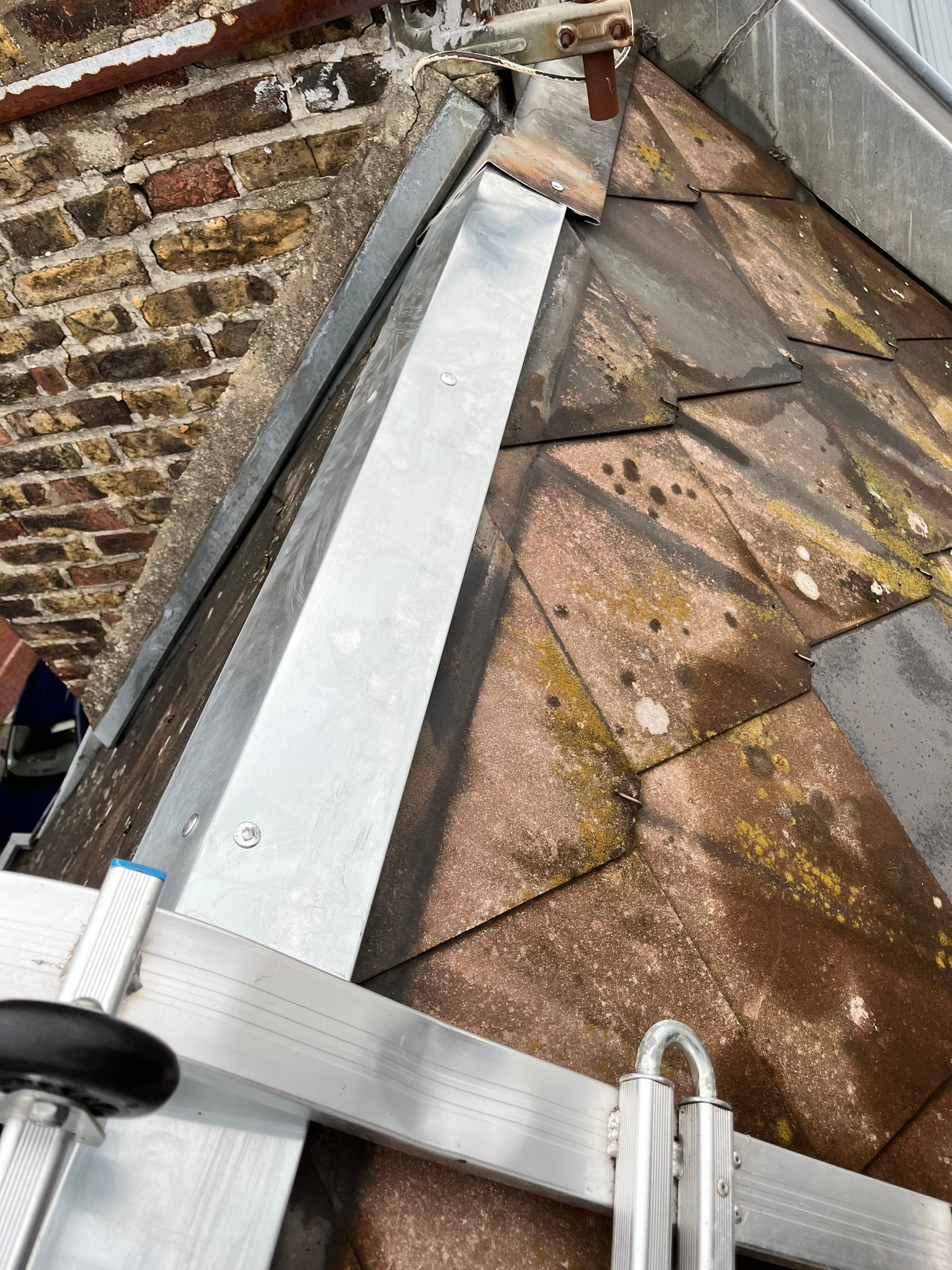 Gros plan sur un angle de toit avec solin métallique et tuiles anciennes et moussues. Échelle visible.