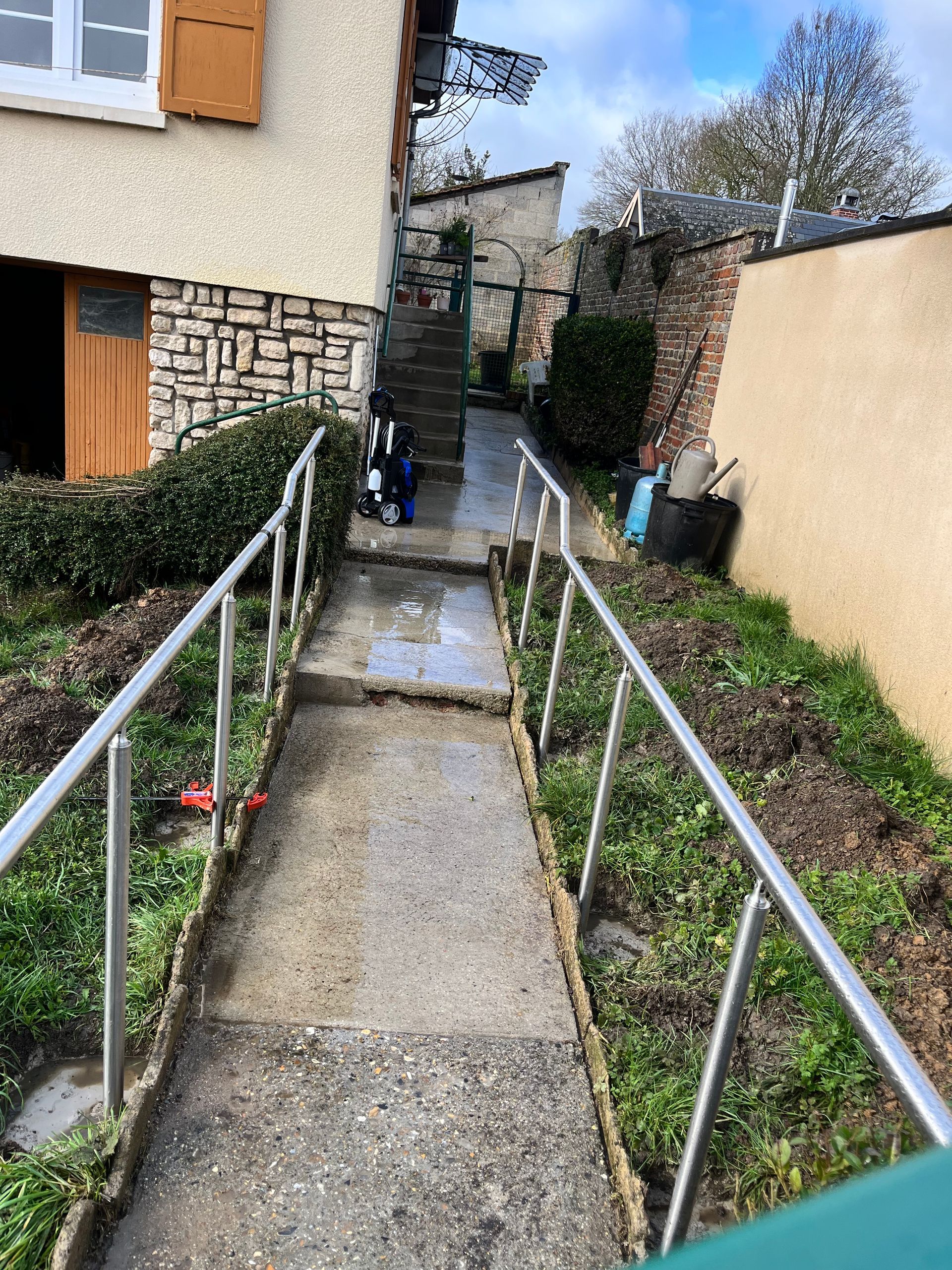 Allée en béton avec rambardes métalliques, montant vers un escalier, à côté d'un bâtiment et de verdure.