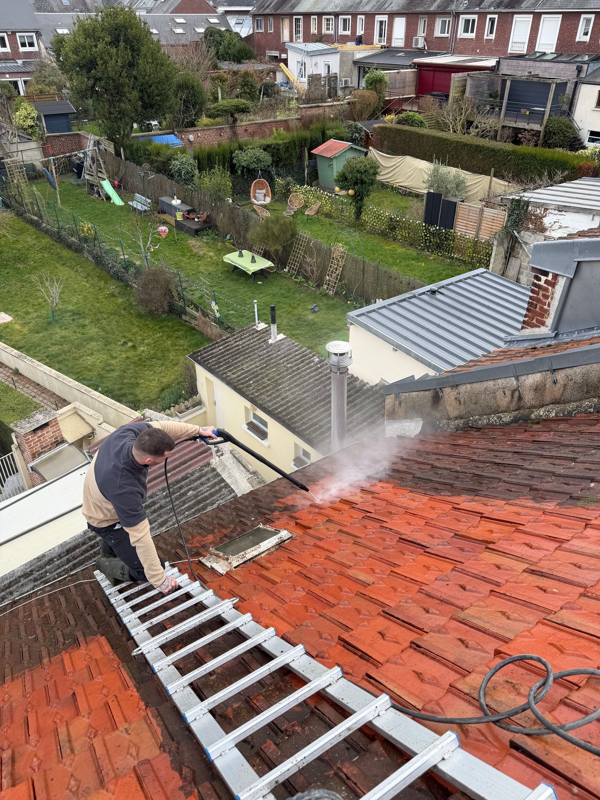 Une personne nettoie à haute pression un toit de tuiles rouges depuis une échelle. En arrière-plan, un quartier résidentiel avec des jardins.