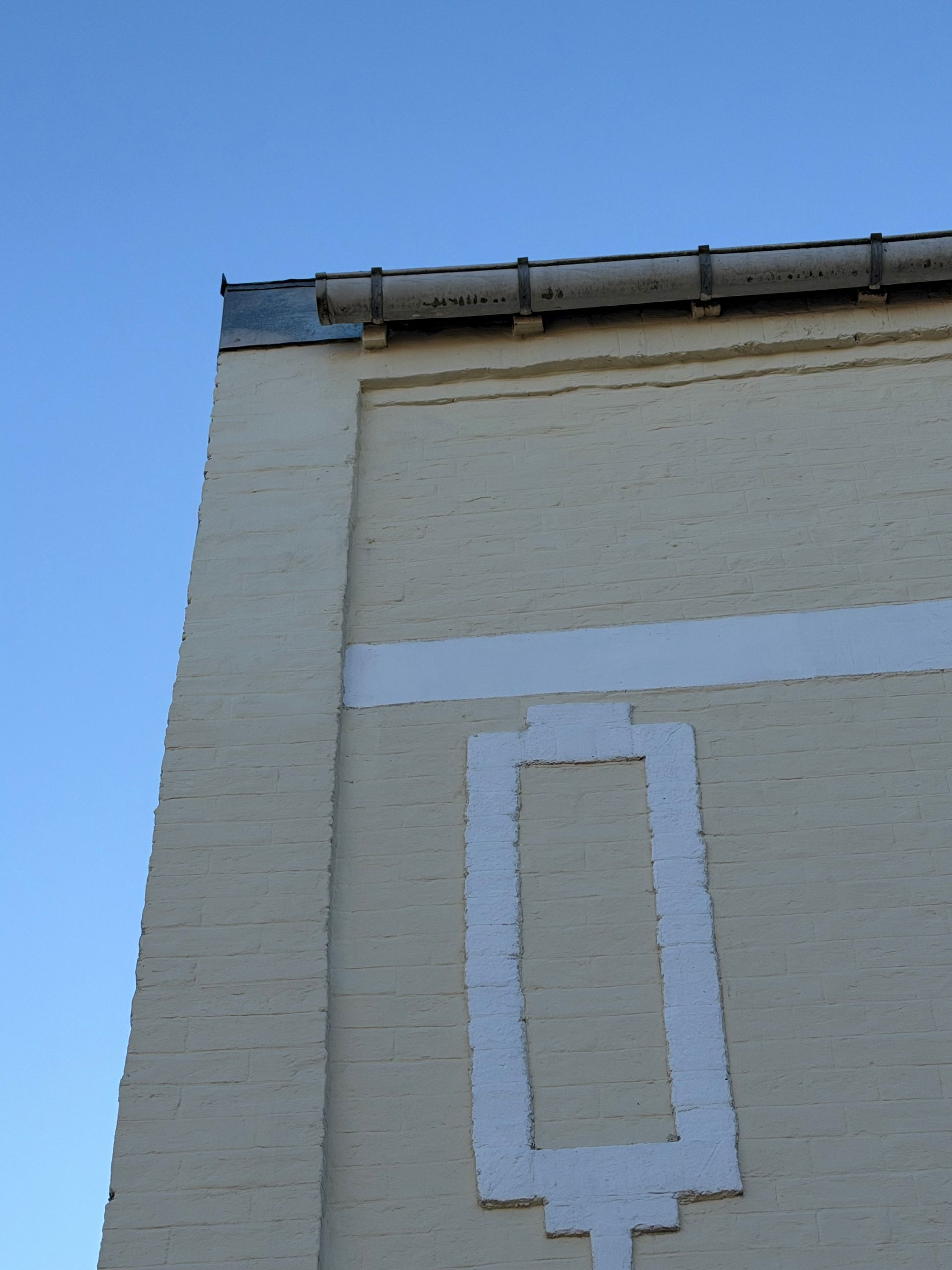 Angle d'un bâtiment en briques blanc cassé, orné d'un rectangle blanc décoratif et se détachant sur un ciel bleu.