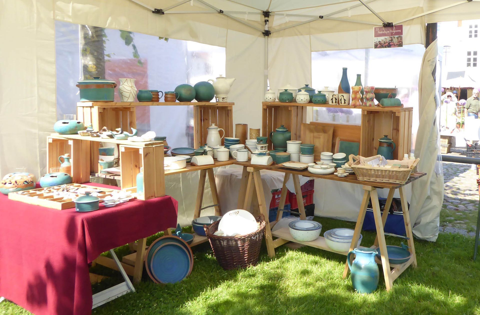 Ein Tisch mit einer roten Tischdecke steht vor einem Zelt voller Töpferwaren.