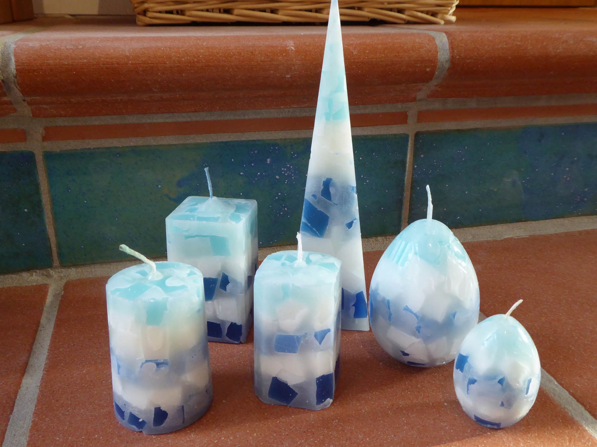 Weiß-blaue Kerzen in Glashaltern, darunter eine hohe, kegelförmige Kerze, auf einer gefliesten Fläche.