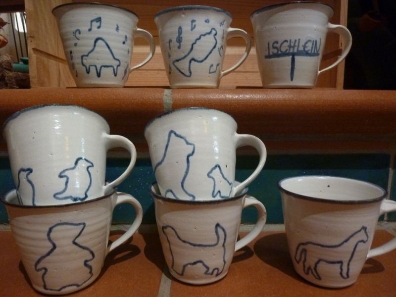 Mehrere Tassen sind übereinander gestapelt auf einem Regal und auf einer steht „lschlei“