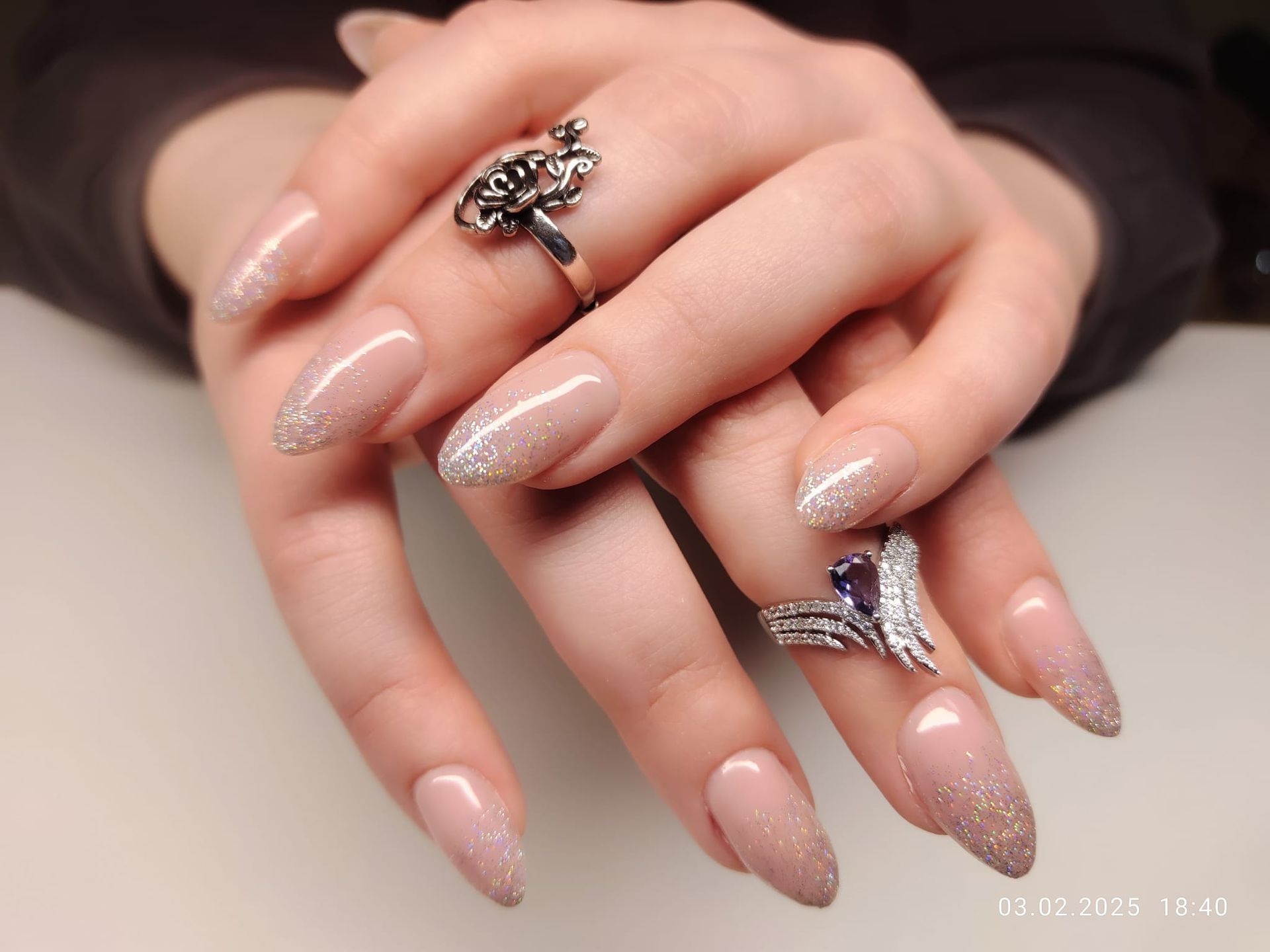 Hände mit langen, mandelförmigen Nägeln. Die Nägel sind nudefarben mit Glitzerspitzen