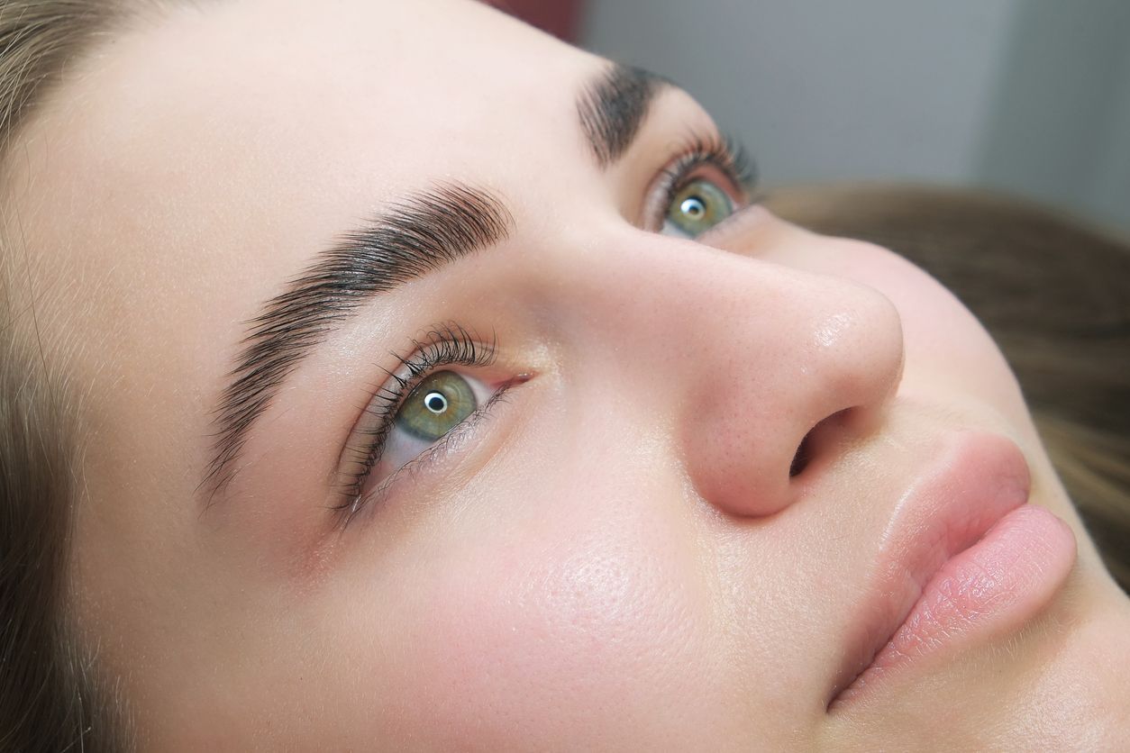 Nahaufnahme eines Gesichts mit grünen Augen, gepflegten Augenbrauen und natürlichem Make-up, das auf einer weichen Oberfläche liegt.