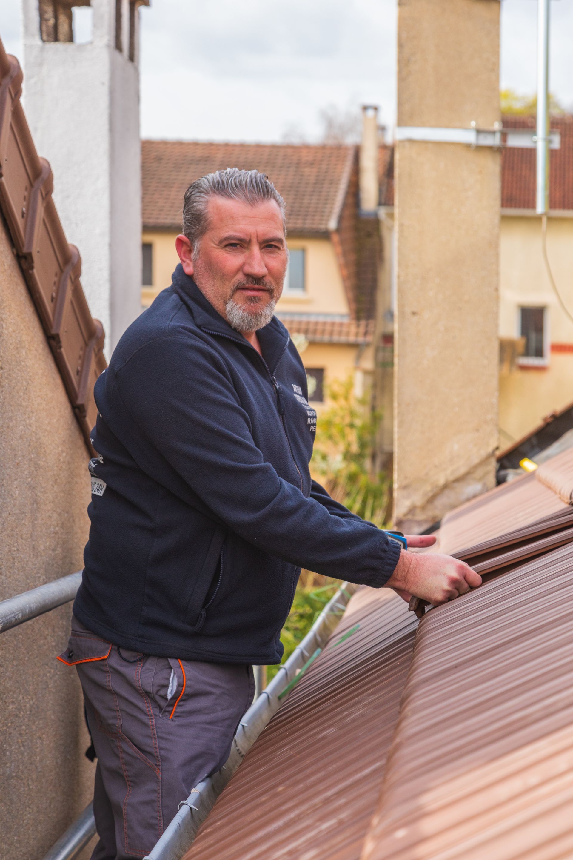 Un couvreur sur un toit de tuiles portant une veste bleue et un pantalon gris.