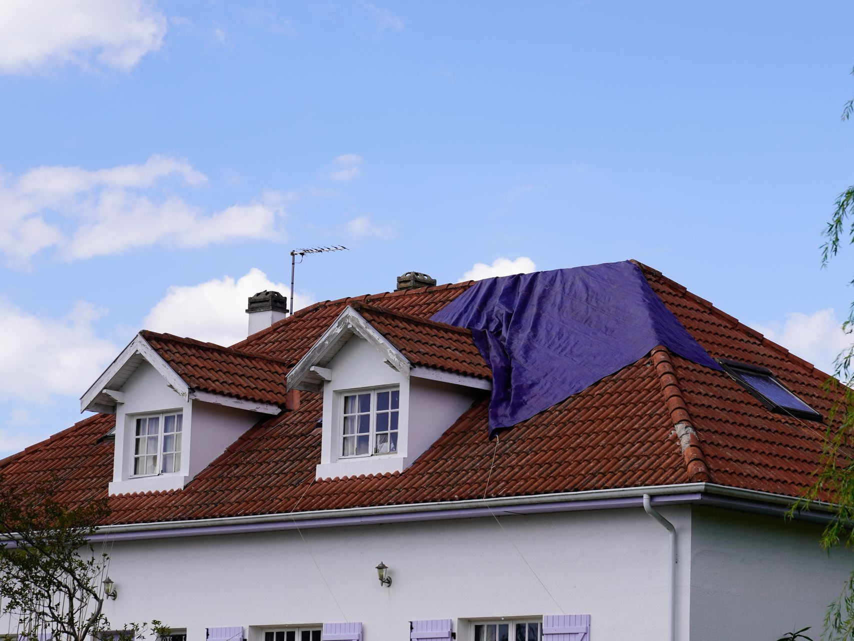Maison avec le toit en tuiles avec une bâche bleue recouvrant une partie endommagée.