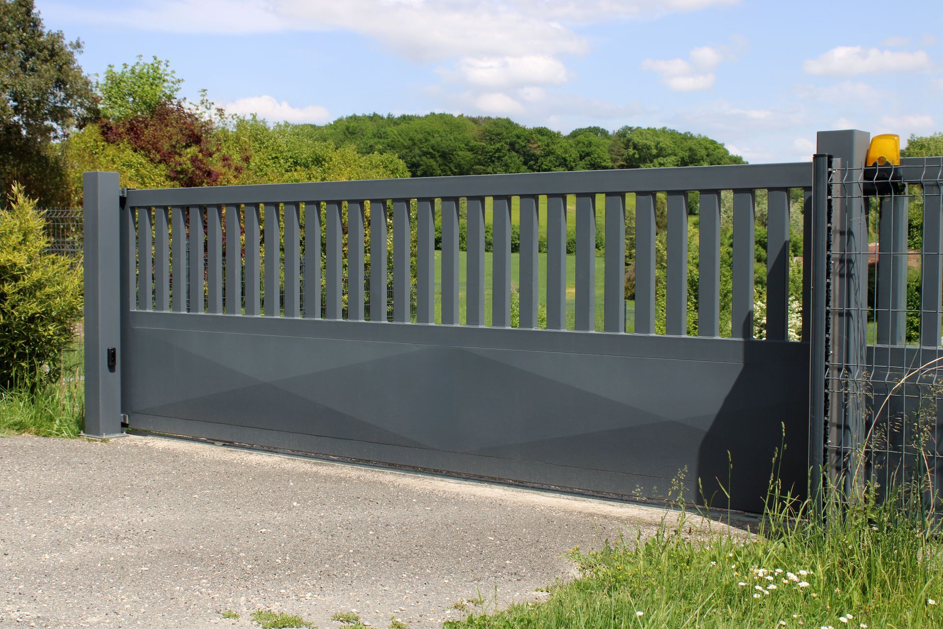 Portail en métal gris sur une allée de gravier, barreaux verticaux en partie supérieure, partie inférieure pleine. Plantes vertes au premier plan.