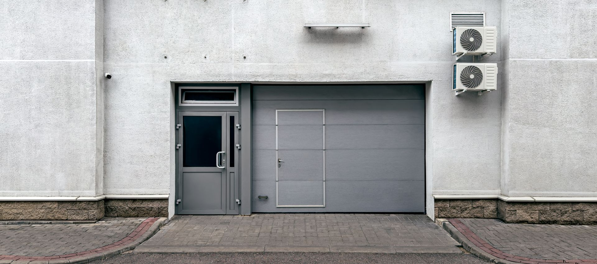 Bâtiment gris avec porte d'entrée, porte de garage et unités de climatisation. Allée en briques devant.