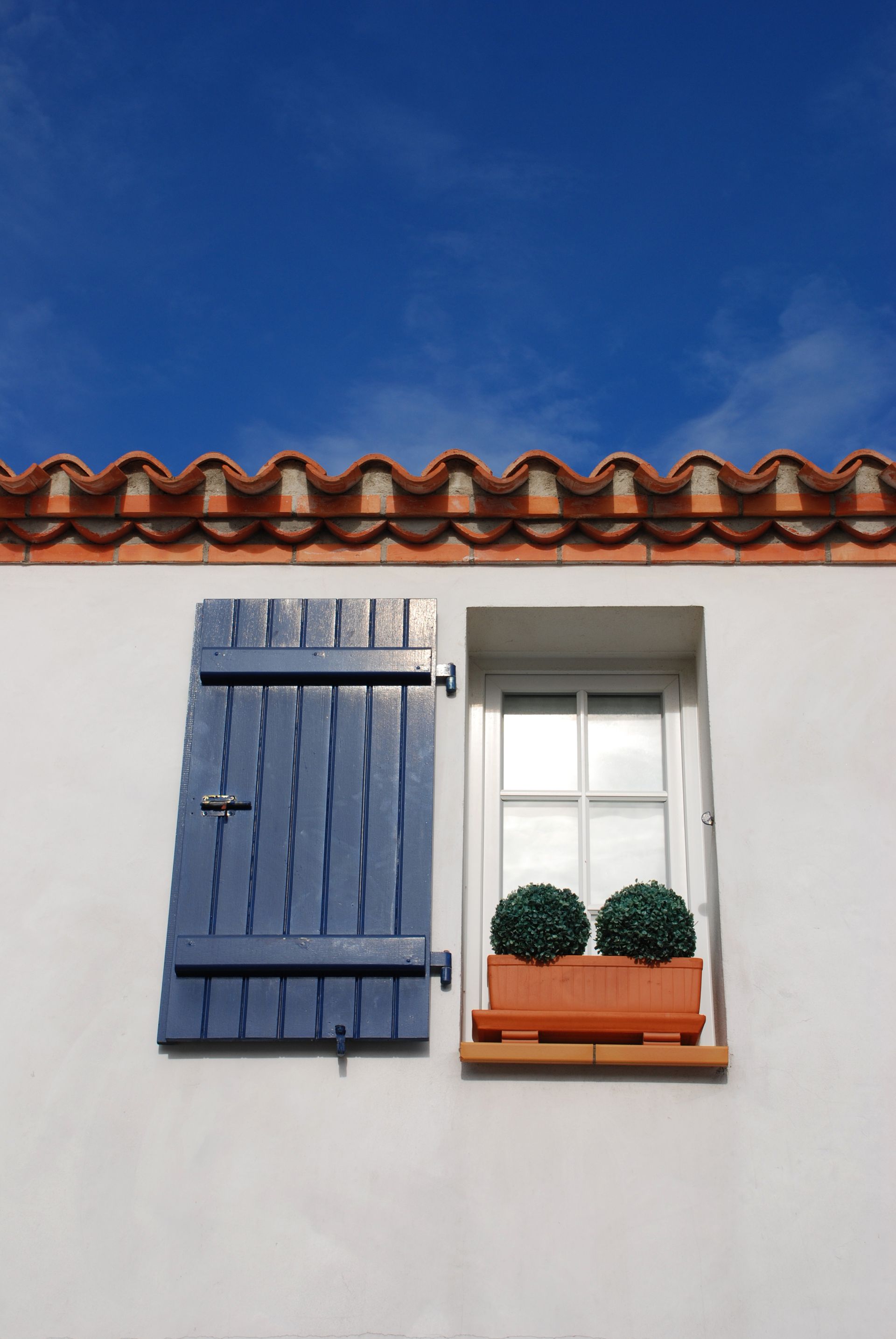 Volet bleu ouvert sur un mur blanc, fenêtre avec deux plantes en pot, toit de tuiles rouges, ciel bleu.
