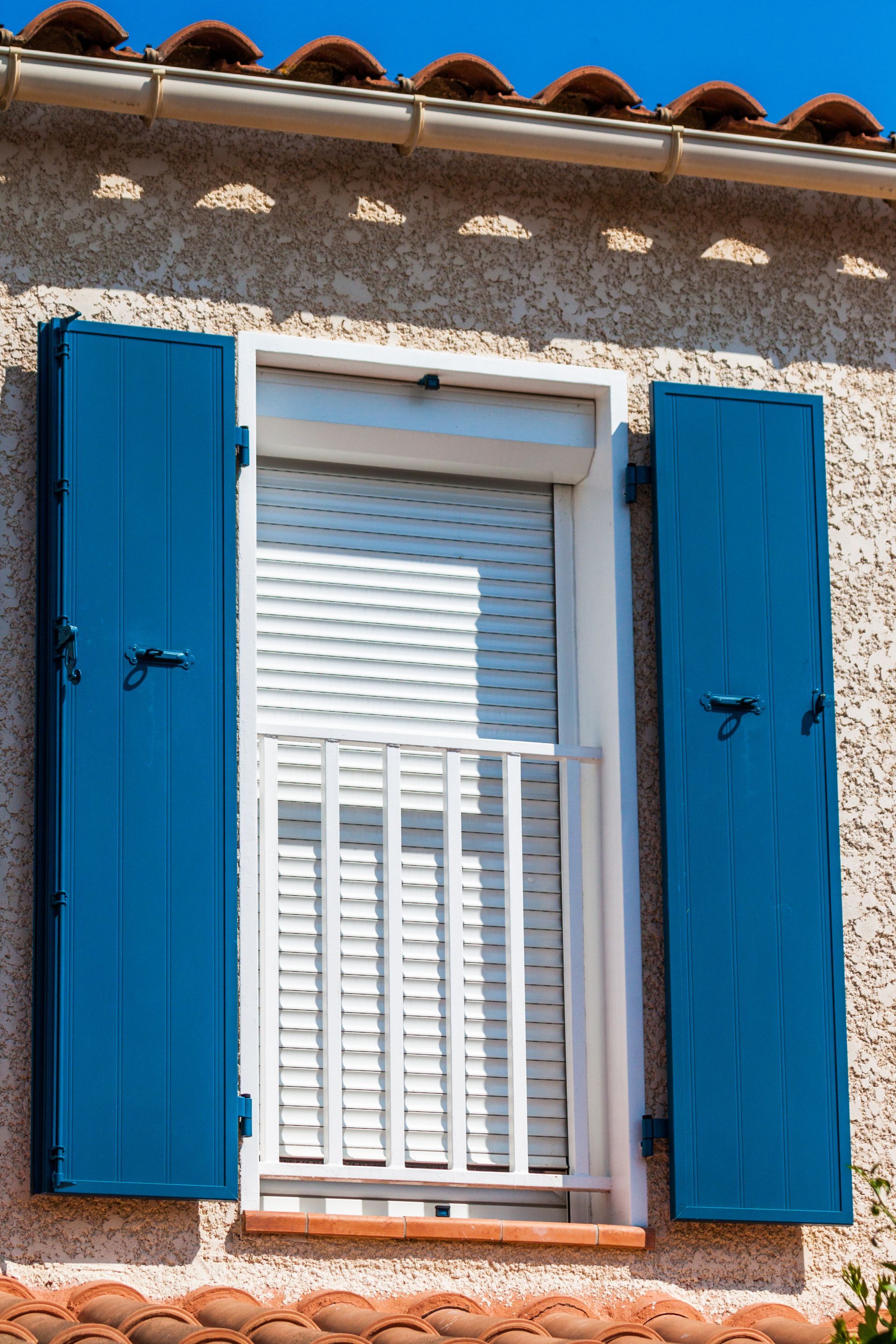 Des volets bleus encadrent une fenêtre blanche dont le volet roulant est fermé et qui donne sur un petit balcon blanc.