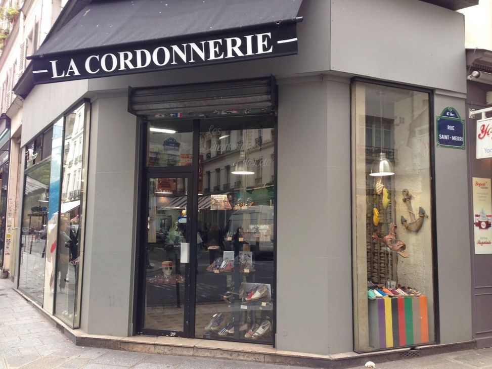 Une boutique d'angle grise avec une façade portant l'inscription LA CORDONNERIE, un auvent noir et des vitrines présentant des chaussures et des sacs.