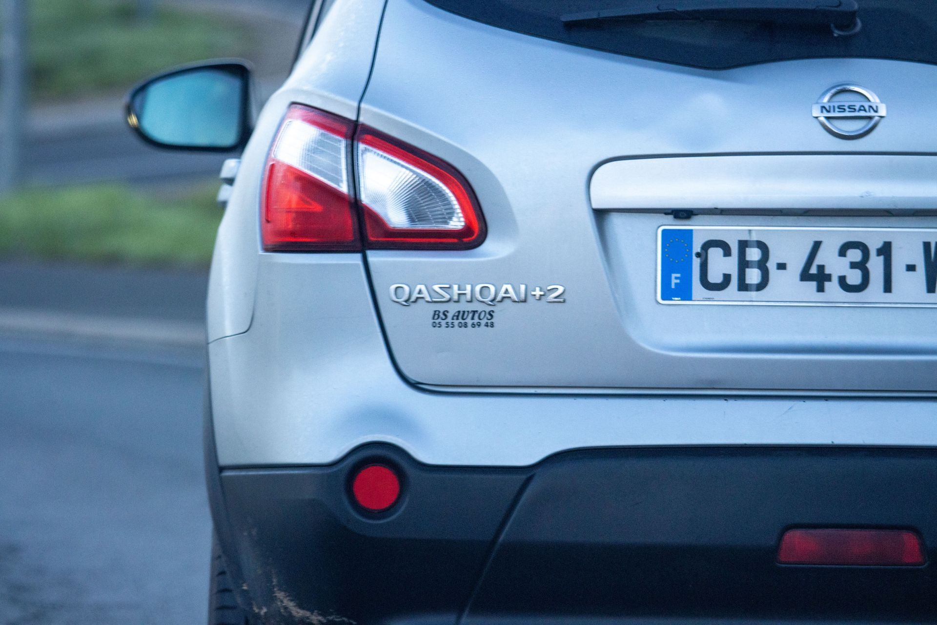Vue arrière d'une Nissan Qashqai+2 argentée, avec un focus sur le feu arrière, le logo du modèle et une partie de la plaque d'immatriculation.