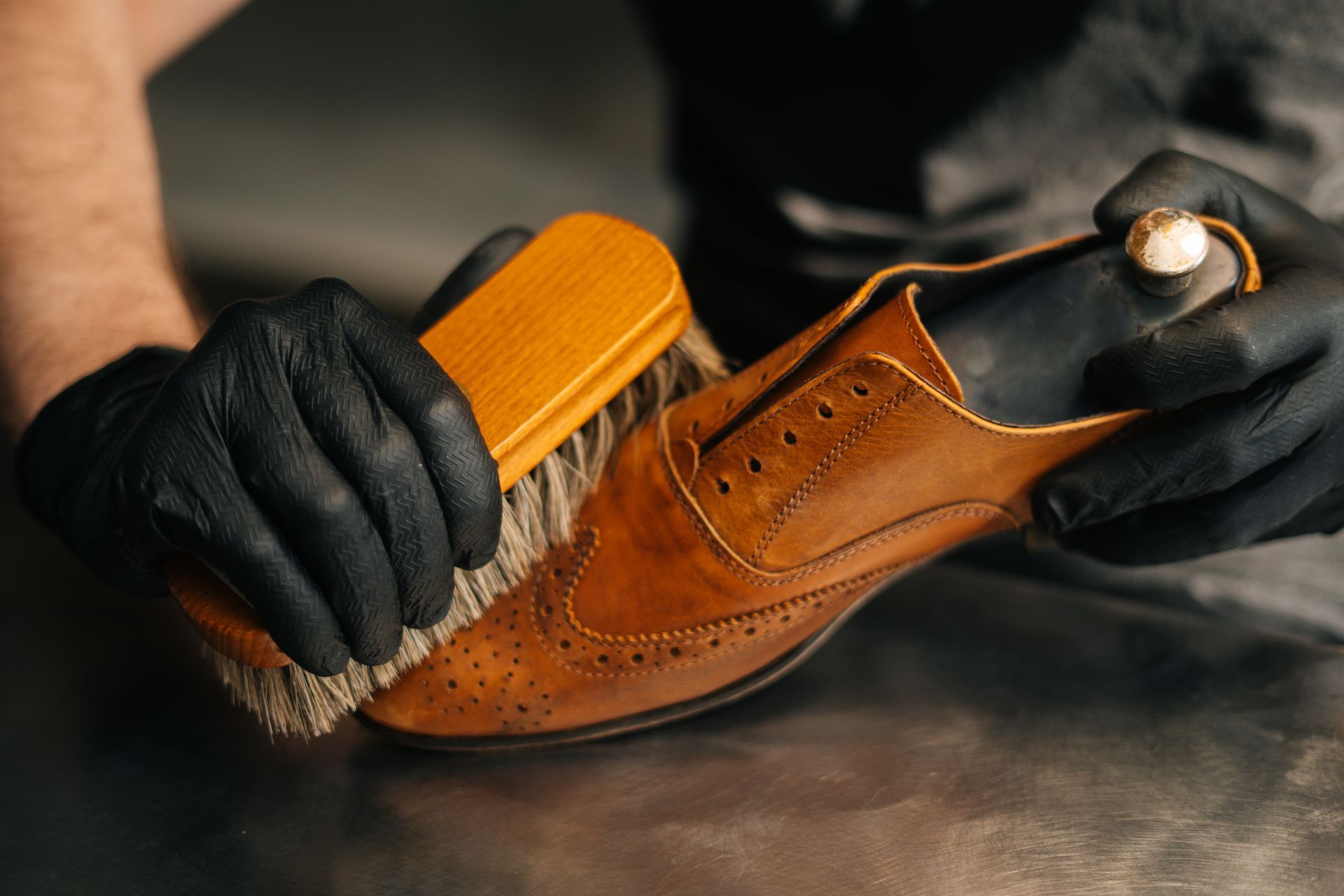 Gros plan sur une personne portant des gants noirs qui utilise une brosse en bois pour cirer une chaussure à bout golf en cuir marron.