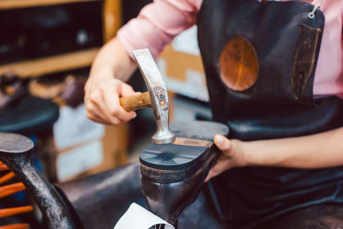 Dans un atelier, une personne portant un tablier en cuir utilise un marteau pour fixer un talon à une chaussure.