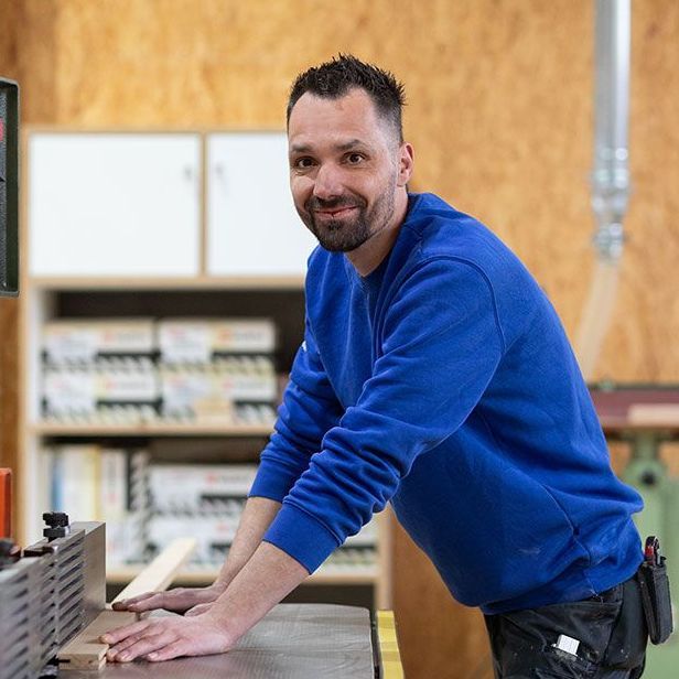 Ein Mann in einem blauen Pullover lehnt an einer Tischkreissäge.