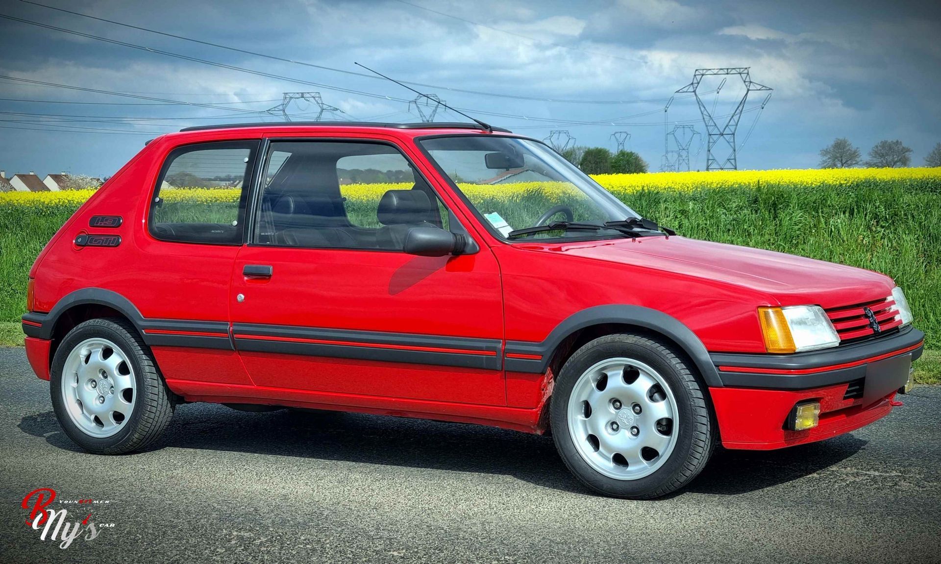 Une Peugeot 205 GTI rouge vif garée sur une route asphaltée devant un champ de fleurs jaunes sous un ciel nuageux.