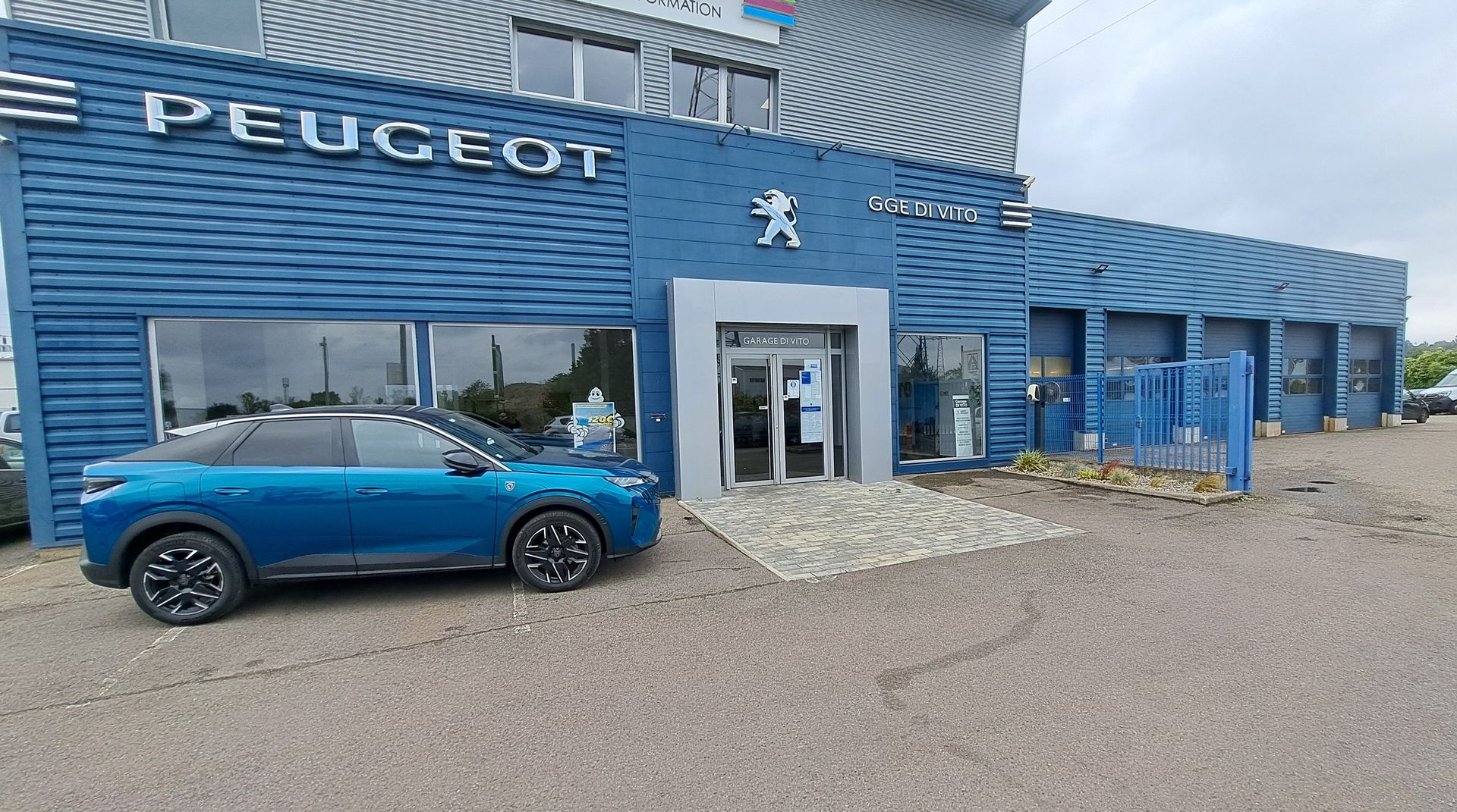 Un bâtiment bleu abritant une concession Peugeot, avec une voiture bleue assortie garée devant, sur un terrain gravillonné.