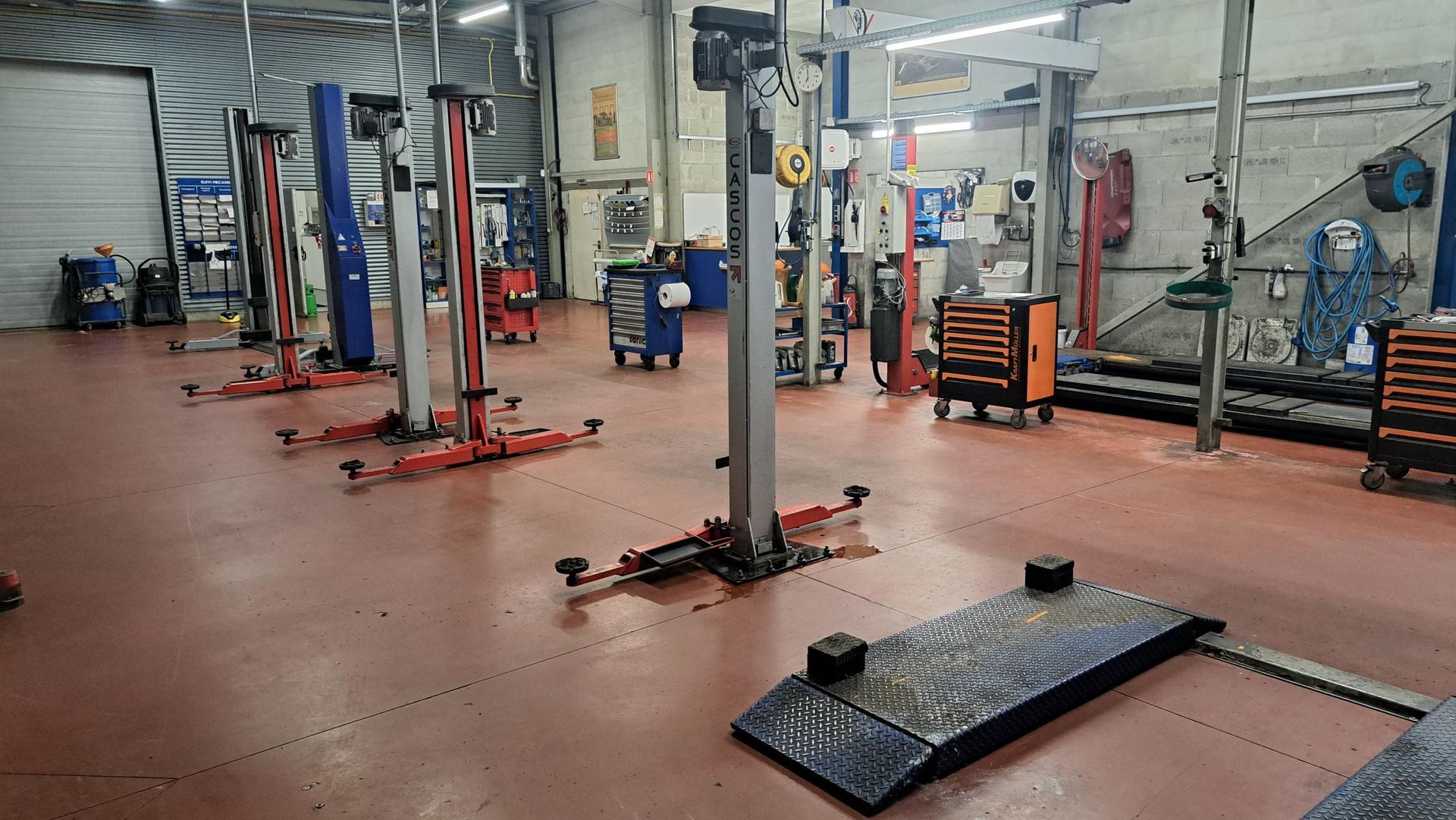 Intérieur d'un atelier de réparation automobile avec plusieurs ponts élévateurs sur un sol rouge.