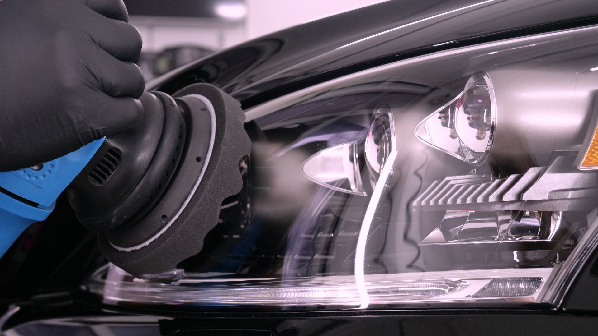 Une personne portant des gants noirs polit un phare de voiture avec une polisseuse électrique.