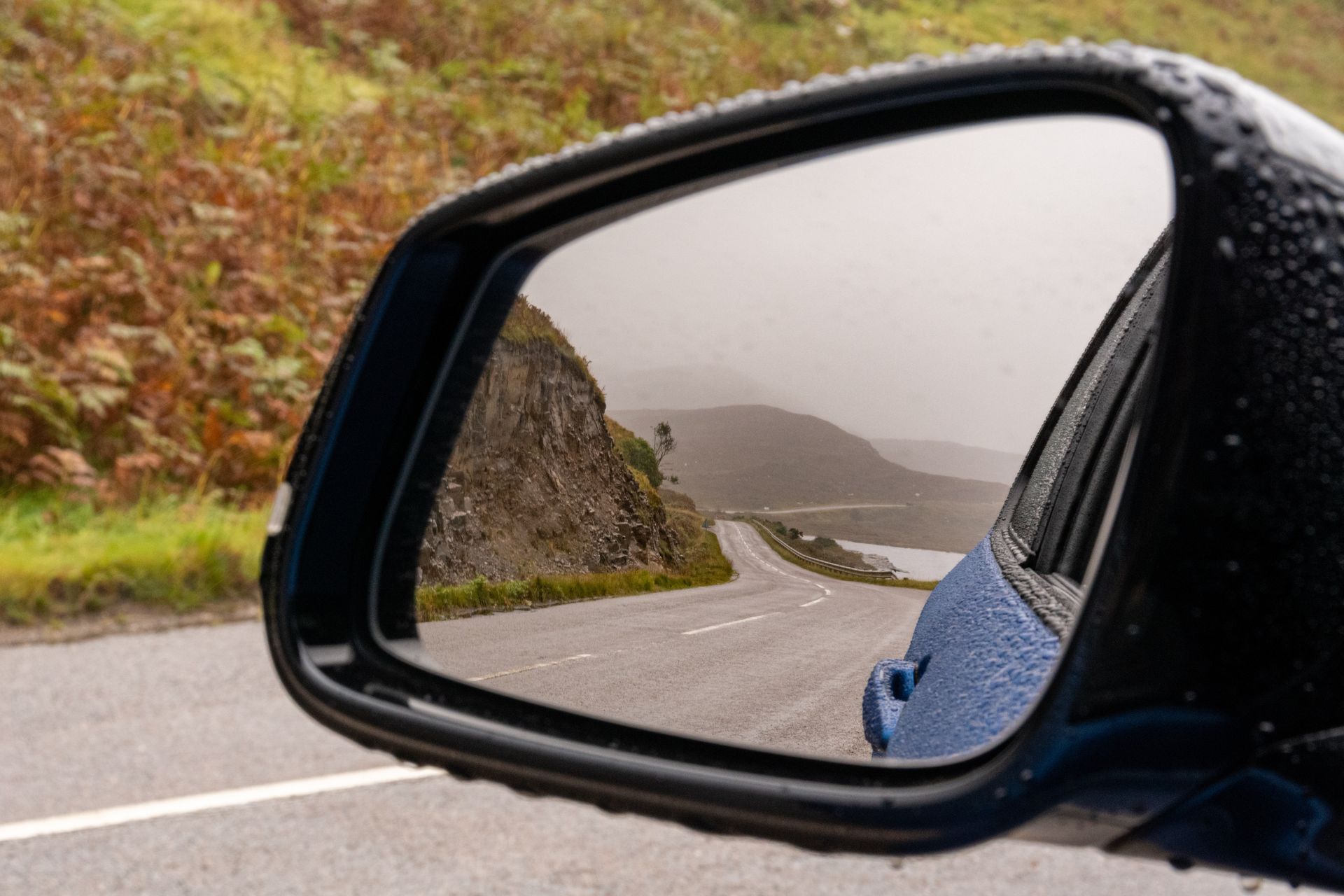 Le rétroviseur d'une voiture reflète une route sinueuse à flanc de colline, avec de l'eau et un ciel nuageux.