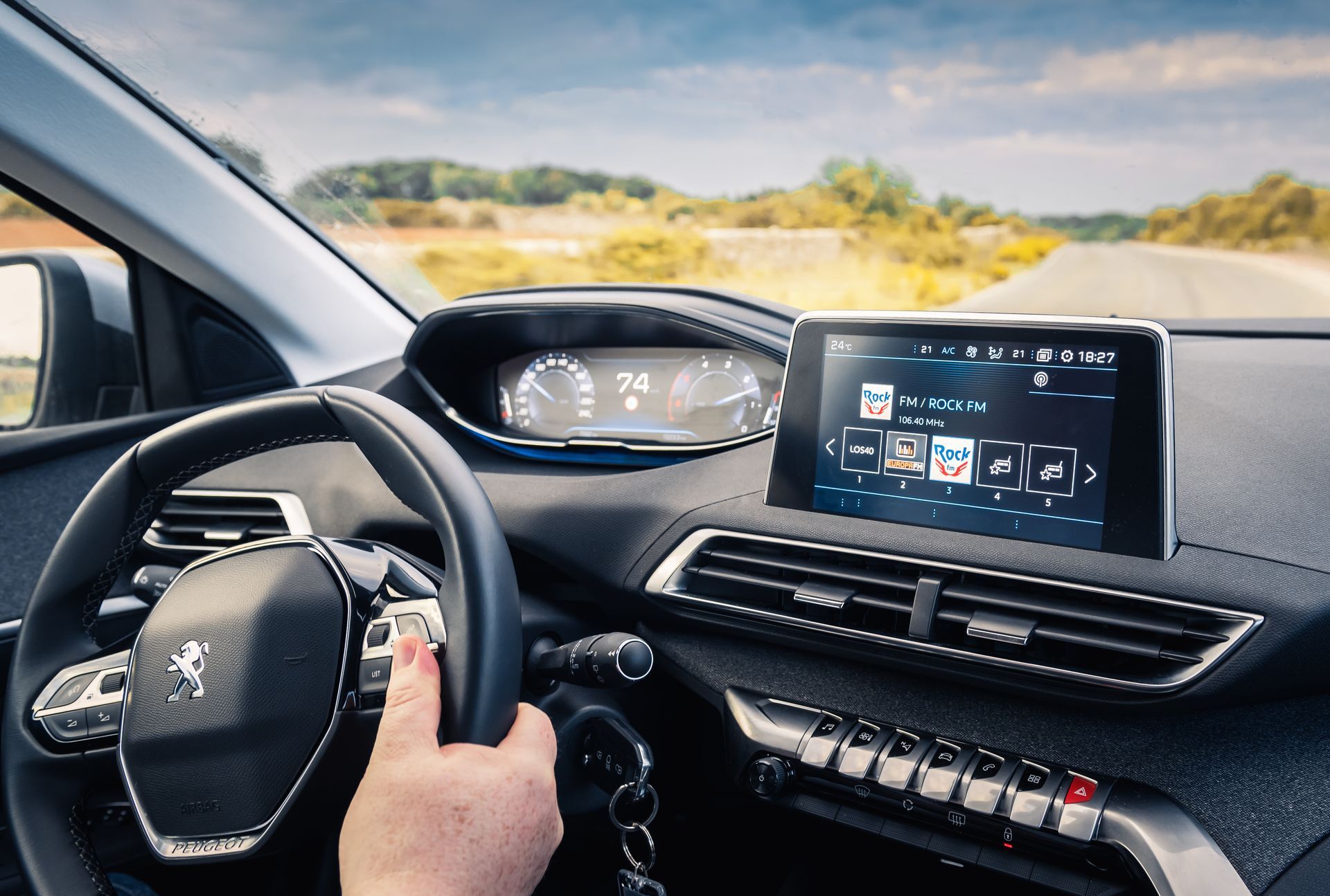 Intérieur d'une voiture ; main sur le volant, tableau de bord avec écran numérique affichant la liste de lecture musicale, route visible à l'extérieur.
