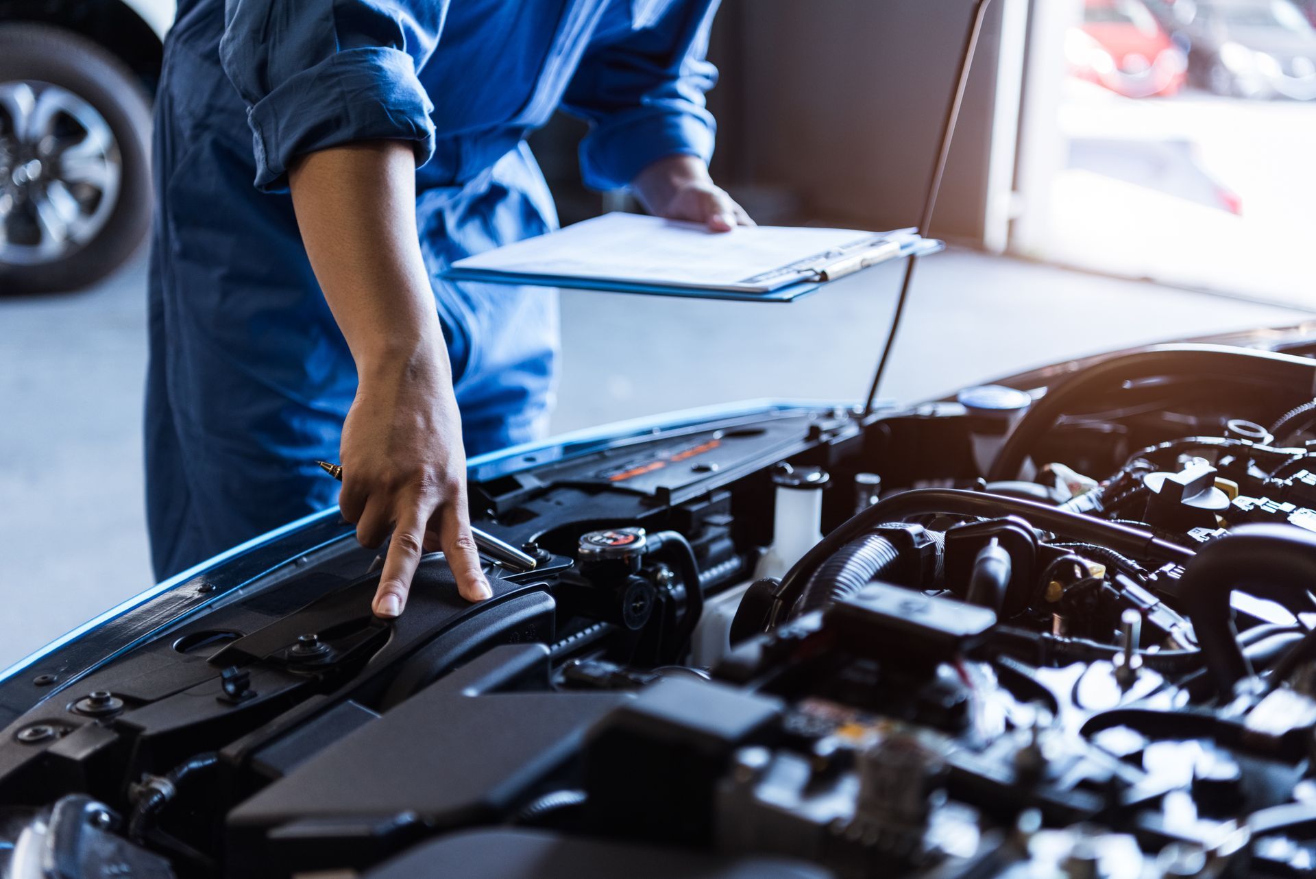 Un mécanicien en combinaison bleue examine le moteur d'une voiture, un bloc-notes à la main, dans un garage.