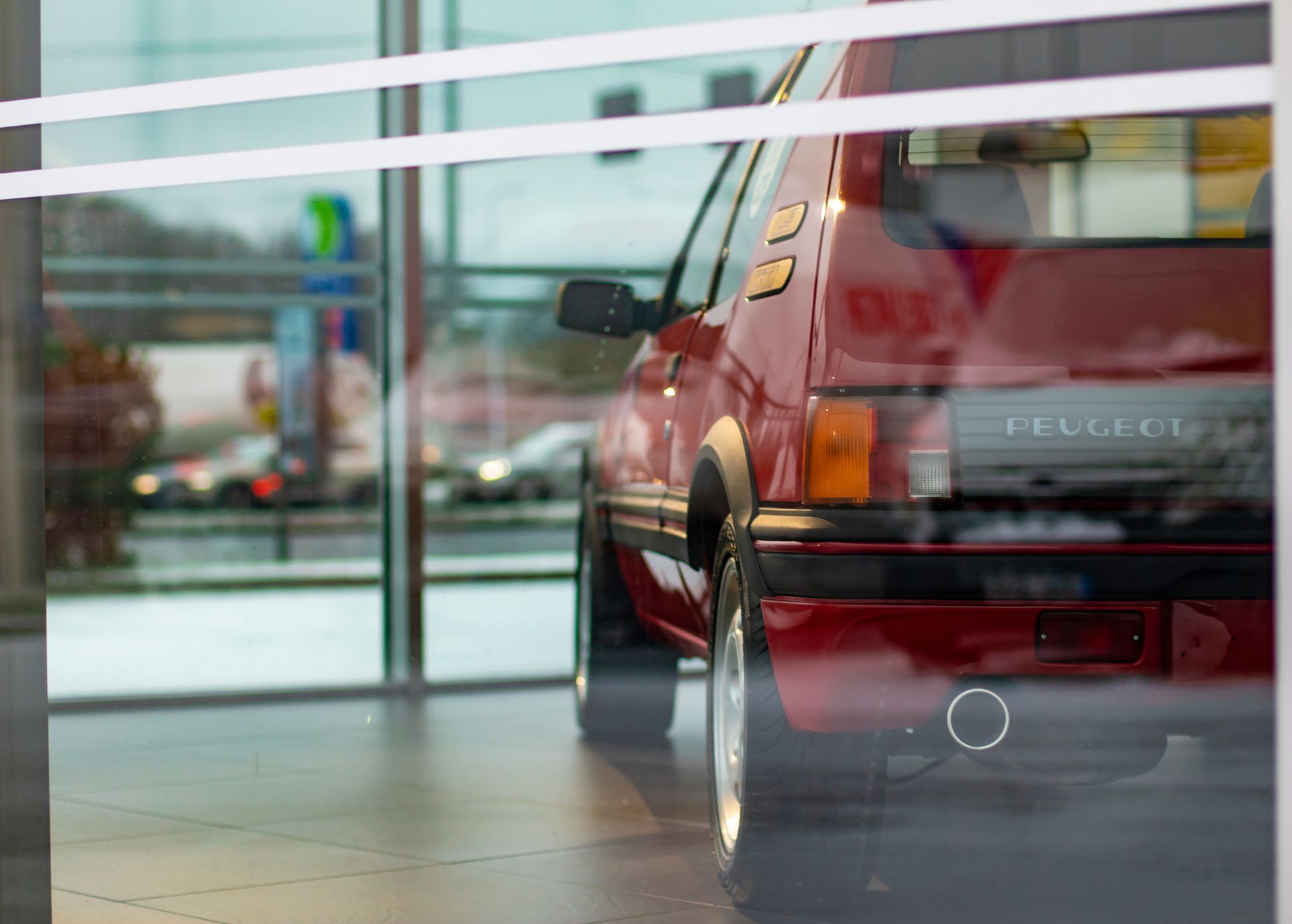 Une Peugeot 205 GTI rouge, garée à l'intérieur, est visible à travers une vitre. L'arrière de la voiture, avec ses feux arrière et le logo Peugeot.