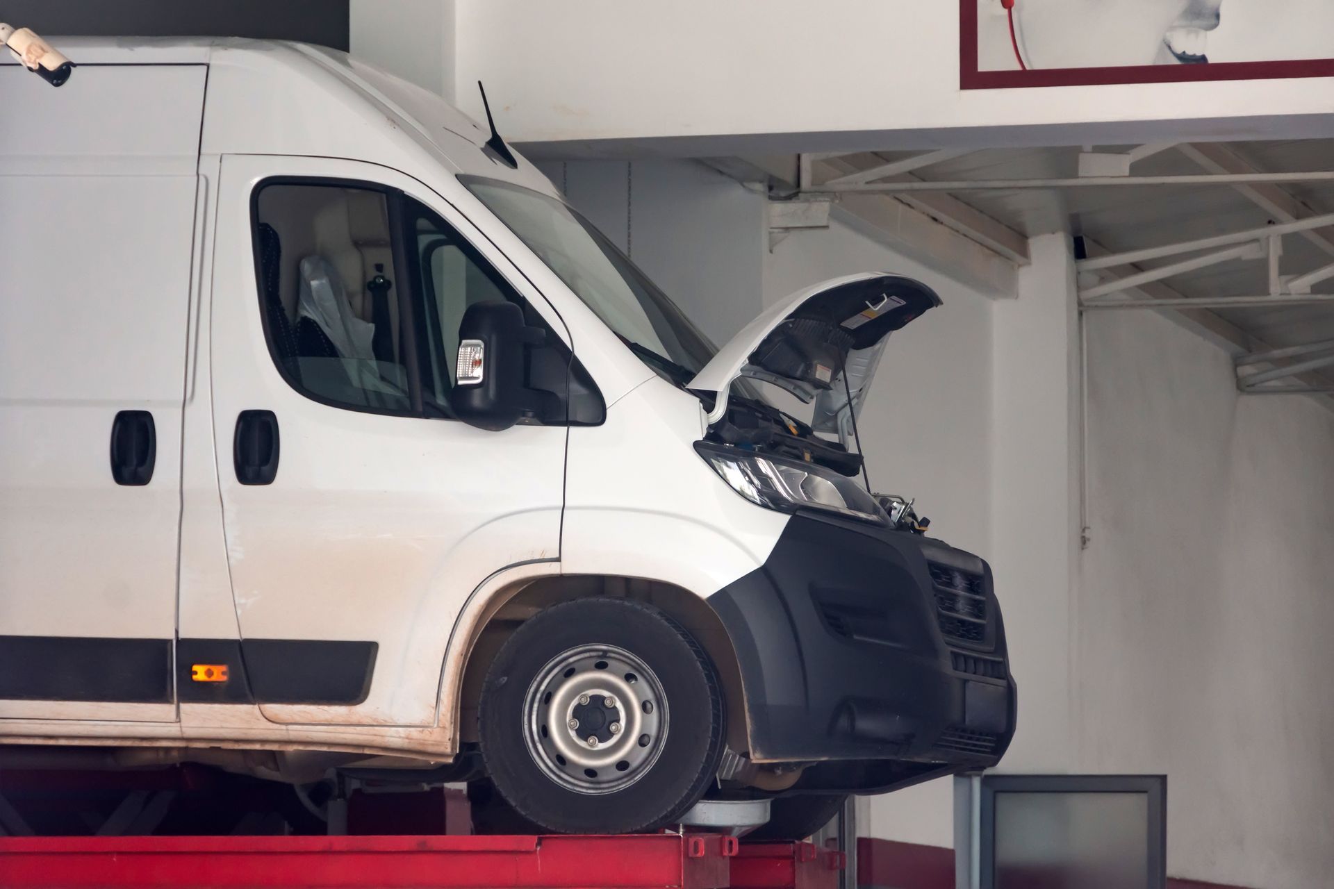Fourgonnette blanche, capot ouvert, sur un pont élévateur rouge dans un atelier de réparation.