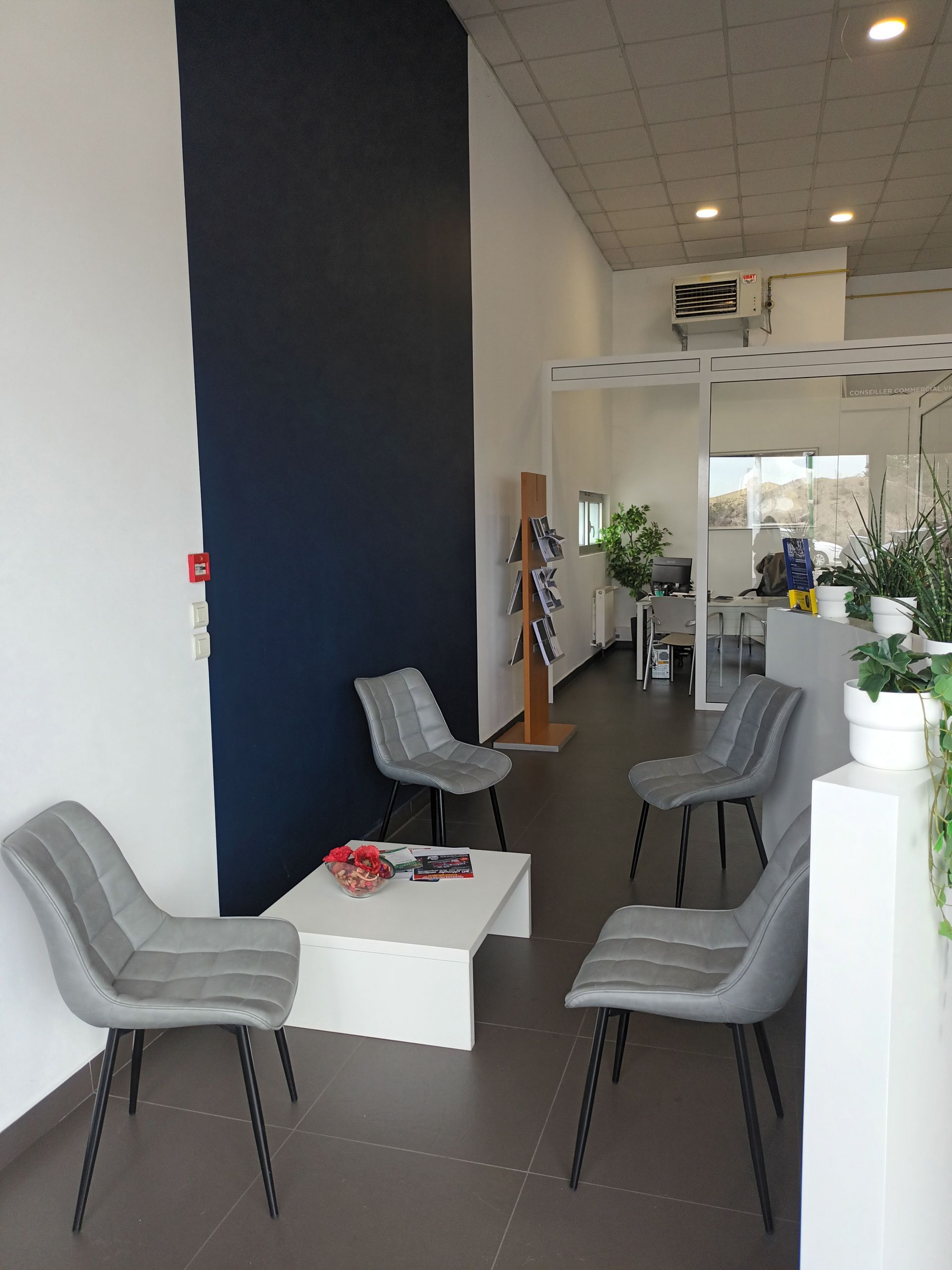 Salle d'attente avec chaises grises, table blanche et mur bleu. Des plantes et un bureau sont visibles en arrière-plan.