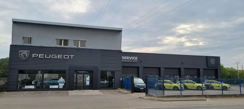 Un bâtiment bleu abritant une concession Peugeot, avec une voiture bleue assortie garée devant, sur un terrain gravillonné.