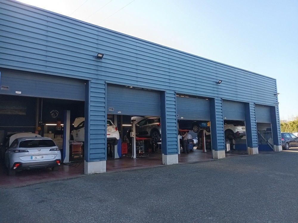 Extérieur d'un atelier de réparation automobile avec trois baies ouvertes, voitures à l'intérieur sur des ponts élévateurs, bardage bleu.