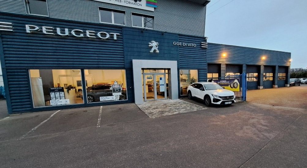 Façade d'une concession Peugeot avec une voiture garée devant. Bâtiment bleu foncé, éclairage nocturne.